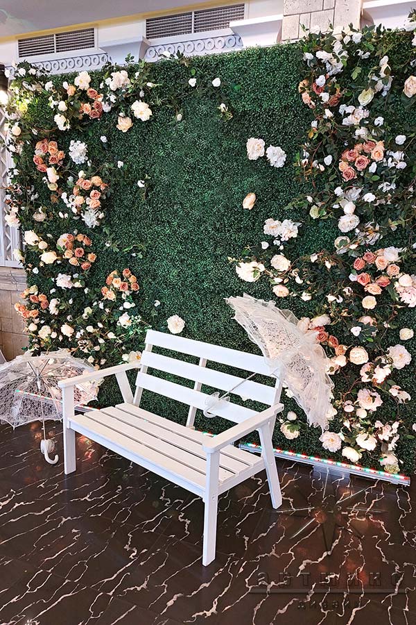 Фотозона из самшита с букетиками роз и белыми зонтиками у скамеечки